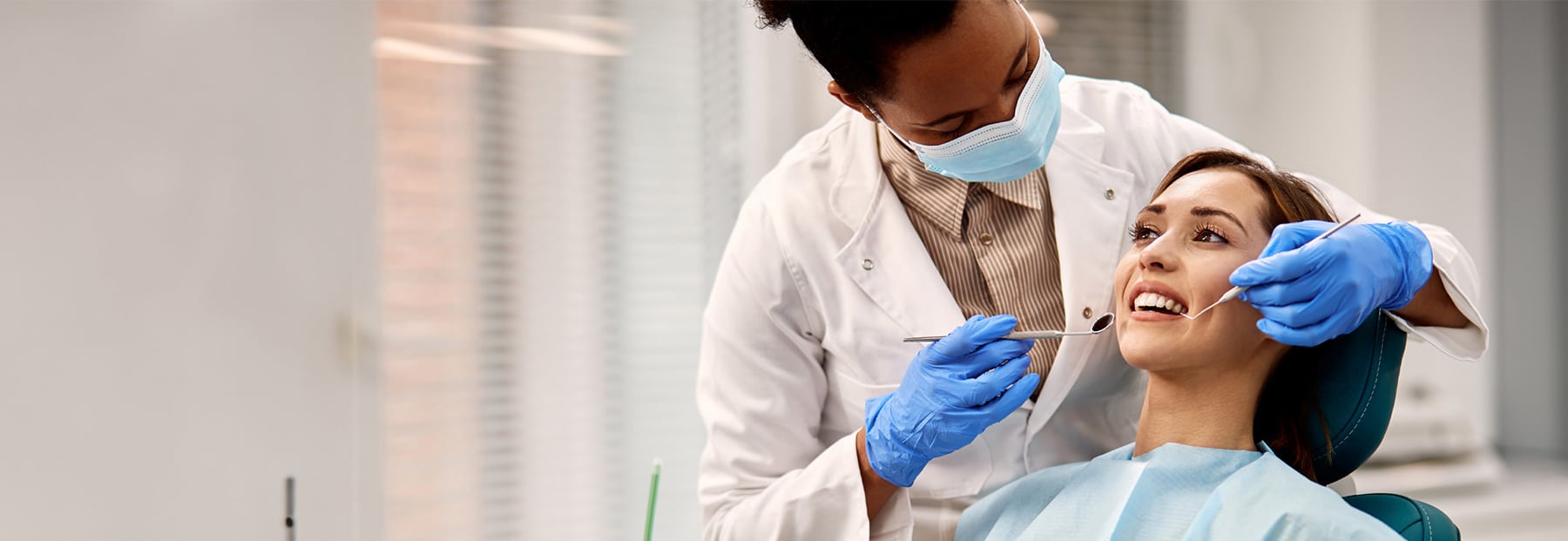 Brown-haired-female in dentist office chair getting serviced by a doctor wearing medical gloves inspecting her teeth.