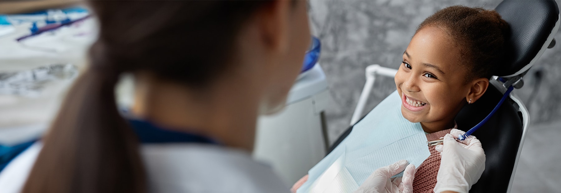 Young patient smiling in dentist office chair as dental assistant puts dental bib around patient.