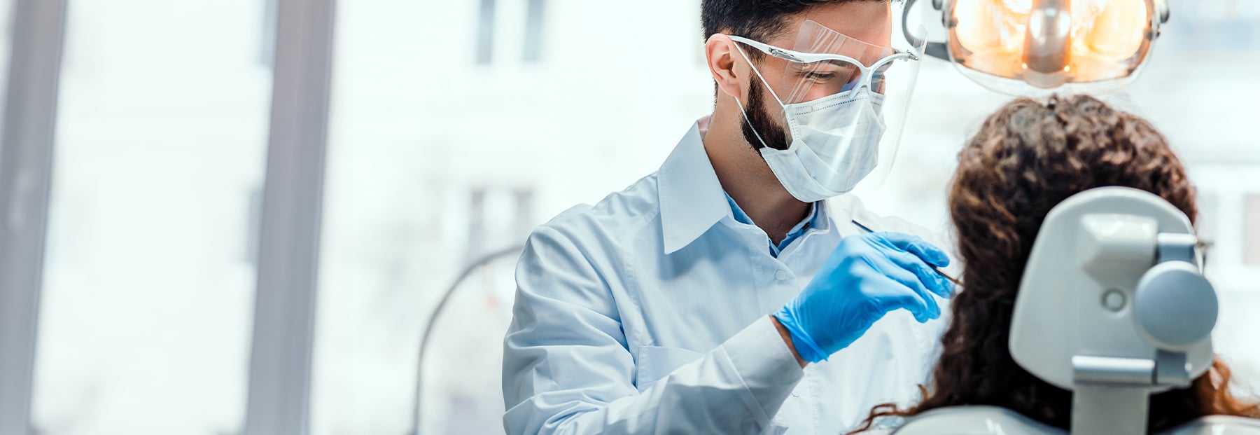 Doctor with eye visor and surgery mask, holding dental tool in front of patient's mouth.