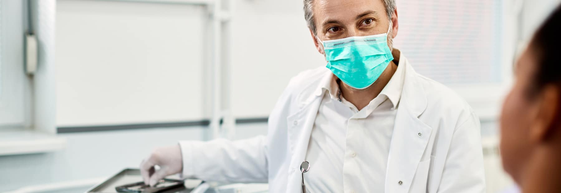 Peppered- hair doctor with surgery mask on and white lab coat, talking to patient.