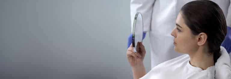 Patient with brown hair holding mirror, looking at her face post surgery.