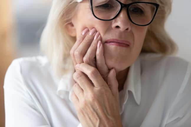 Close up of elderly woman with black glasses holding jaw from tooth pain.